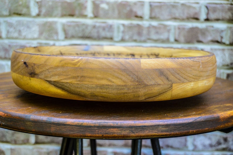 Puede incluir: Un cuenco de madera hecho a mano con un dise&ntilde;o ancho y poco profundo. El cuenco est&aacute; hecho de varias piezas de madera, mostrando patrones de vetas naturales y tonos c&aacute;lidos. Se encuentra sobre un taburete de madera oscura, con una pared de ladrillo al fondo.