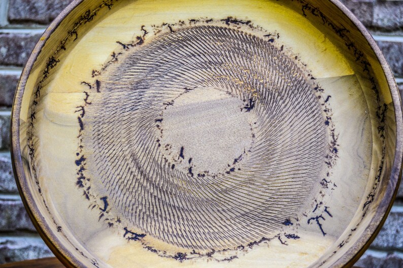 Puede incluir: Una bandeja redonda de madera con un interior de color claro y un patr&oacute;n oscuro en espiral en el centro. El borde es de un tono marr&oacute;n m&aacute;s oscuro. El dise&ntilde;o de la bandeja presenta una serie de l&iacute;neas conc&eacute;ntricas, creando un efecto texturizado.