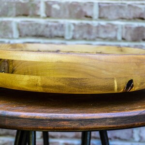 Puede incluir: Un cuenco de madera hecho a mano con un dise&ntilde;o ancho y poco profundo. El cuenco est&aacute; hecho de madera de color claro con vetas y nudos visibles, y est&aacute; sobre una mesa marr&oacute;n oscuro. El fondo es una pared de ladrillo.