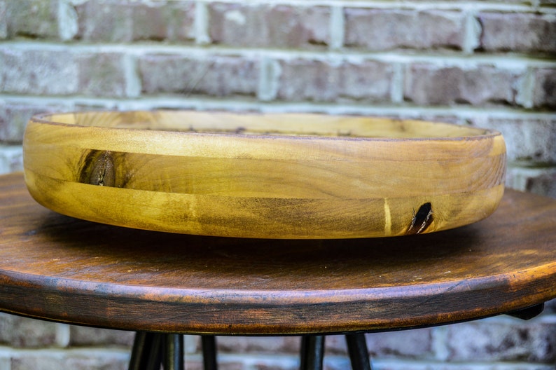 Puede incluir: Un cuenco de madera hecho a mano con un dise&ntilde;o ancho y poco profundo. El cuenco est&aacute; hecho de madera de color claro con vetas y nudos visibles, y est&aacute; sobre una mesa marr&oacute;n oscuro. El fondo es una pared de ladrillo.