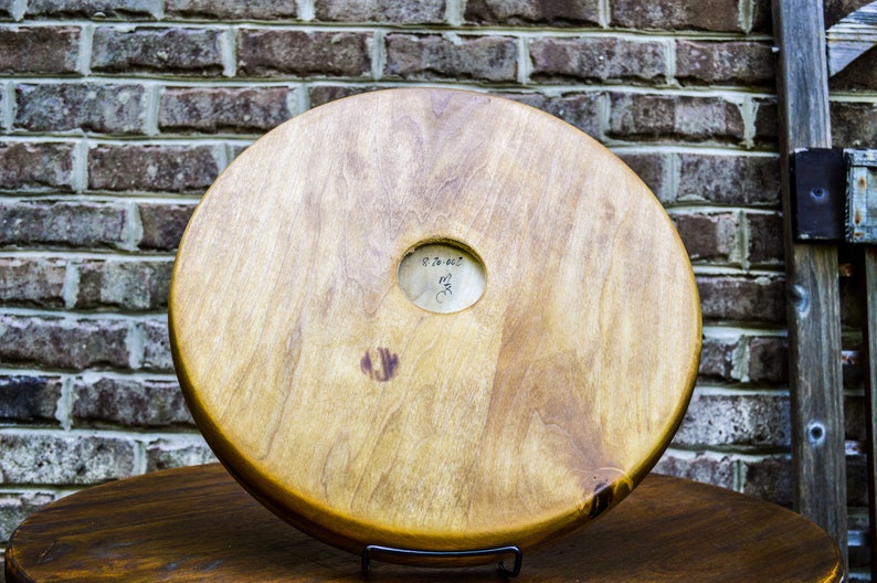 Puede incluir: Objeto de madera redondo y claro, con un agujero central y veta visible. Se muestra sobre una superficie marr&oacute;n oscuro, con una pared de ladrillos al fondo. Una peque&ntilde;a marca circular est&aacute; cerca del centro.