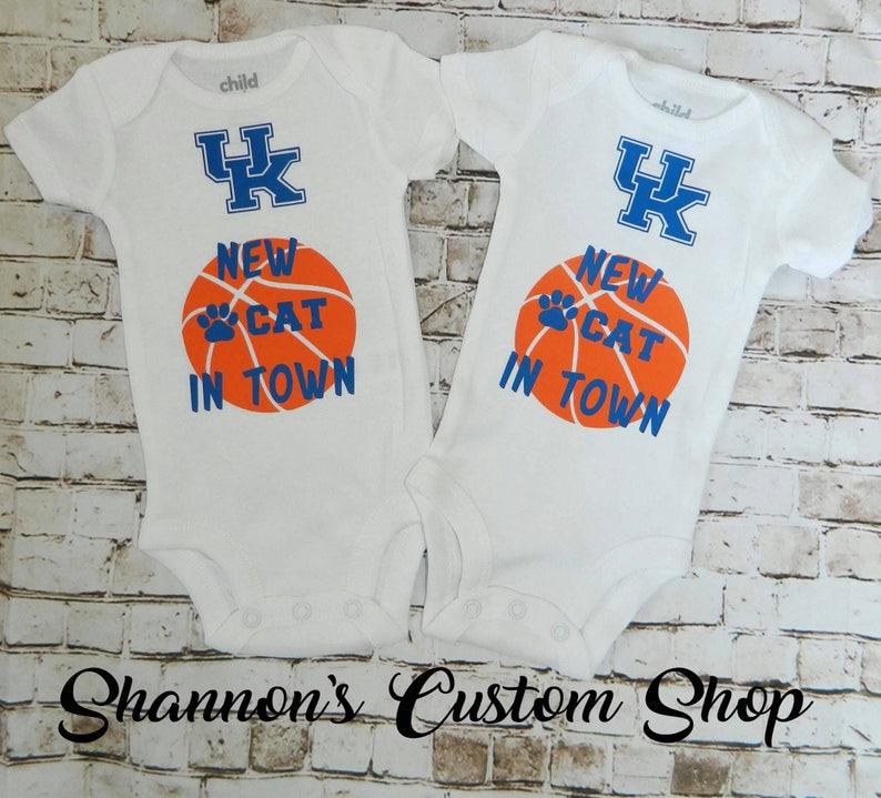 May include: Two white baby onesies with blue and orange text and a basketball graphic. The text reads "New Cat in Town" and the onesies have a blue "UK" logo.