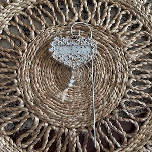 May include: A heart-shaped ornament with a textured, speckled white and brown finish, attached to a silver-toned metal wire stake. A small jute string hangs from the bottom of the heart, featuring two white beads and a small silver-toned spacer bead.