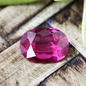 May include: A vibrant pink oval-shaped gemstone with a faceted cut. The gemstone is resting on a wooden surface.