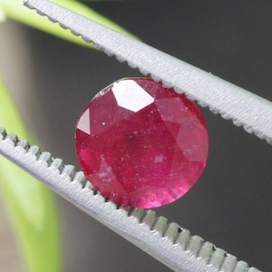 Puede incluir: Una gema facetada, redonda, de color rojo intenso, es sostenida por unas pinzas plateadas. La gema tiene un corte brillante, que refleja la luz. En el fondo se ve follaje verde, con un fondo gris oscuro. La gema es el punto focal de la imagen.