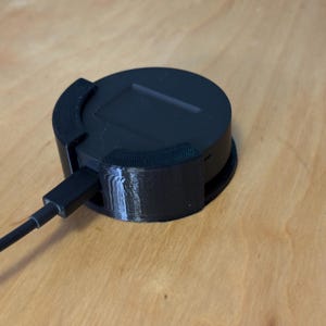 May include: A black, circular electronic device with a rectangular top and a black charging cable plugged into the side. The device is on a light brown wooden surface.