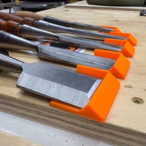 May include: A collection of woodworking chisels with wooden handles and silver blades, each fitted with bright orange blade protectors. The tools are arranged on a light-colored wooden surface, suggesting a workshop setting.