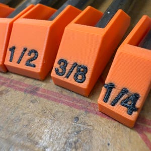 May include: A set of bright orange 3D-printed plastic tool organizers featuring black embossed text labels. The visible labels clearly display the fractional measurements 1/2, 3/8, and 1/4, indicating the specific size of the tool held within each slot.