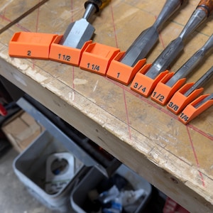 May include: A set of bright orange plastic chisel guards, each featuring a black printed size label ranging from 1/4 inch to 2 inches. These protective covers are fitted over the metal blades of various woodworking chisels with wooden handles.