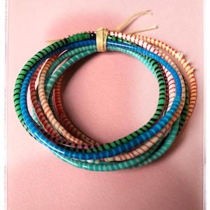 Peut inclure: Un ensemble de bracelets rayés colorés, verts, bleus, rouges et oranges, maintenus ensemble par une bande de couleur naturelle. Les bracelets sont disposés en cercle sur un fond rose.