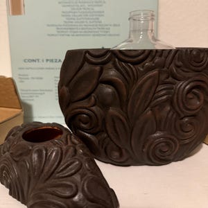 May include: A brown, carved ceramic fragrance holder with a clear glass bottle. The holder features a detailed floral design. A smaller, matching piece sits in front. The background includes a box with text.