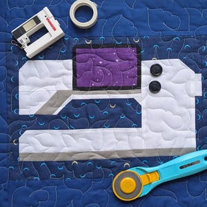May include: A white and gray sewing machine quilt block with a purple star-filled screen and two black buttons. The block is on a blue fabric background with a white and gray quilting pattern. A rotary cutter and a roll of tape are also in the image.