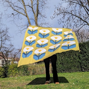 Op de afbeelding: Een gele stoffen deken met een herhalend patroon van witte bloemen met blauwe stelen. De deken wordt vastgehouden door een persoon in een park.