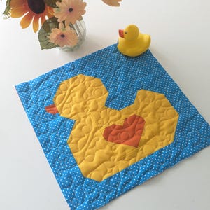 May include: A handmade, quilted baby mat featuring a large yellow duck with an orange heart on a blue polka dot background. A yellow rubber duck toy sits on the mat. The mat is approximately 12 inches square.