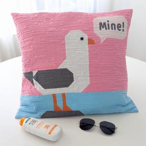 May include: Pink decorative pillow with a cartoon seagull design. The seagull is white, gray, and orange, with the word "Mine!" in a speech bubble. A bottle of sunscreen and sunglasses are in the foreground.
