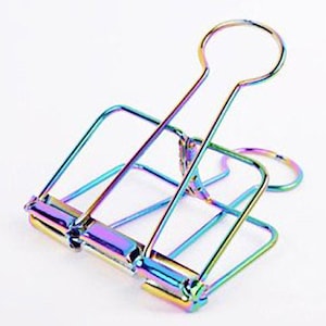 Op de afbeelding: Een regenboogkleurige metalen binderclip met een afgeronde bovenste handgreep. De clip is ontworpen om papier bij elkaar te houden.