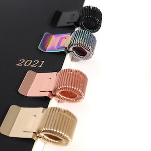 May include: Four metal page markers in different colors: black, iridescent, rose gold, and gold. Each marker has a spring-loaded coil and a flat base for holding pages.