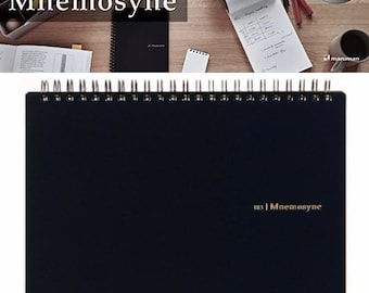 Op de afbeelding: Een zwart spiraalnotitieblok met gouden letters "183 | Mnemosyne". Het notitieblok ligt op een houten oppervlak.