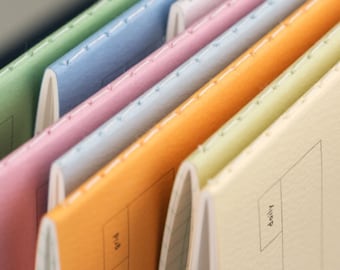 Puede incluir: Una pila de cuadernos coloridos con bordes cosidos. Los cuadernos están etiquetados con "Qty" y "P/B".