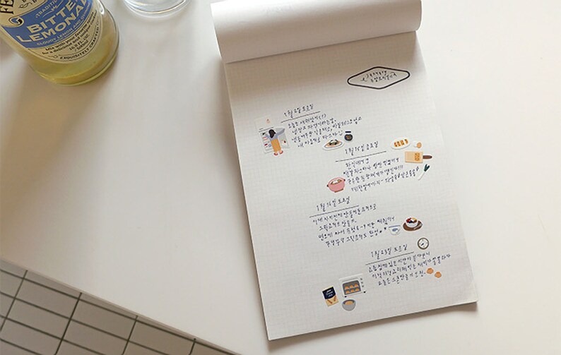Pu&ograve; includere: Un quaderno bianco con un motivo a griglia e note scritte a mano in coreano. Il quaderno ha un piccolo adesivo a forma di diamante con la parola "Planner" scritta su di esso. Ci sono anche diversi piccoli adesivi colorati che raffigurano cibo e persone.