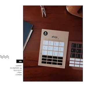 May include: Two sheets of clear calendar stickers with black and white text. The stickers are on a brown surface. The text on the stickers reads "plain deco +" and includes the months of the year.