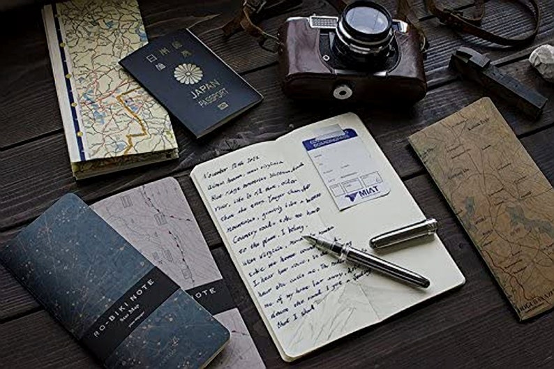 May include: A flat lay of travel essentials including a passport, a notebook, a pen, and a vintage camera. The notebook has handwritten notes and a boarding pass. The passport is for Japan.