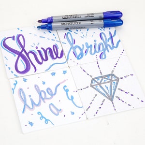 May include: Four white ceramic tiles with hand-painted designs in shades of blue, purple, and silver. The tiles feature the words "Shine bright" and "like a" with a diamond design. Two silver and blue markers are lying on the table.