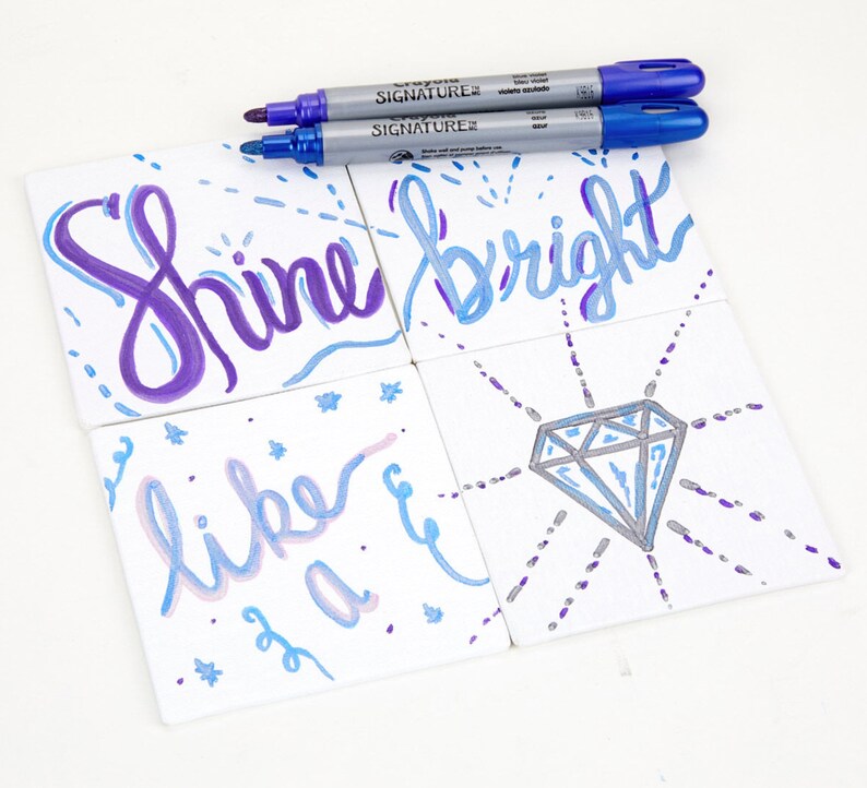 May include: Four white ceramic tiles with hand-painted designs in shades of blue, purple, and silver. The tiles feature the words "Shine bright" and "like a" with a diamond design. Two silver and blue markers are lying on the table.