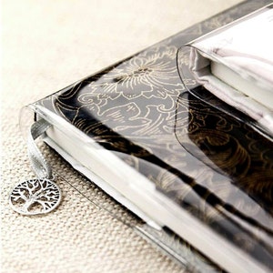 May include: A black and gold patterned journal with a clear plastic cover and a silver tree charm attached to a ribbon bookmark.