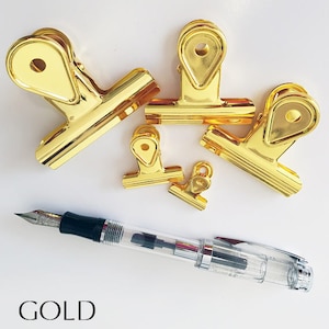May include: Five gold binder clips of different sizes, with a clear and black fountain pen on a white background. The word "GOLD" is written in black at the bottom of the image.