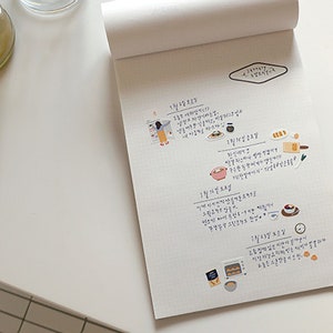 Pu&ograve; includere: Un quaderno bianco con un motivo a griglia e note scritte a mano in coreano. Il quaderno ha un piccolo adesivo a forma di diamante con la parola "Planner" scritta su di esso. Ci sono anche diversi piccoli adesivi colorati che raffigurano cibo e persone.