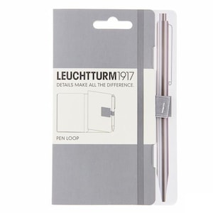 Puede incluir: Un embalaje gris y blanco para un cuaderno Leuchtturm1917 con un bucle para bolígrafo y un bolígrafo plateado. El embalaje tiene el texto "LEUCHTTURM1917" y "DETAILS MAKE ALL THE DIFFERENCE."