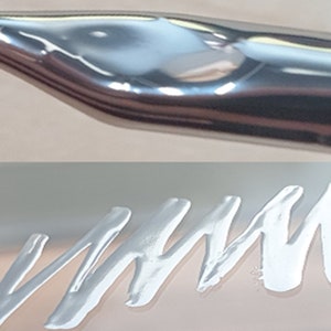 Könnte beinhalten: Nahaufnahme einer silbernen Kalligraphie-Feder mit einer spitzen Spitze. Die Feder liegt auf einer weißen Oberfläche mit einer silbernen, wellenförmigen Linie, die darauf gezeichnet ist.