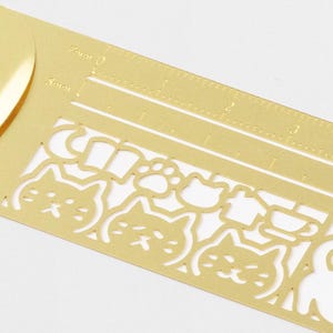 May include: A gold-coloured metal ruler featuring cut-out cat faces, paw prints, and other shapes. The ruler has measurement markings in centimetres and millimetres. Ideal for crafting and scrapbooking.