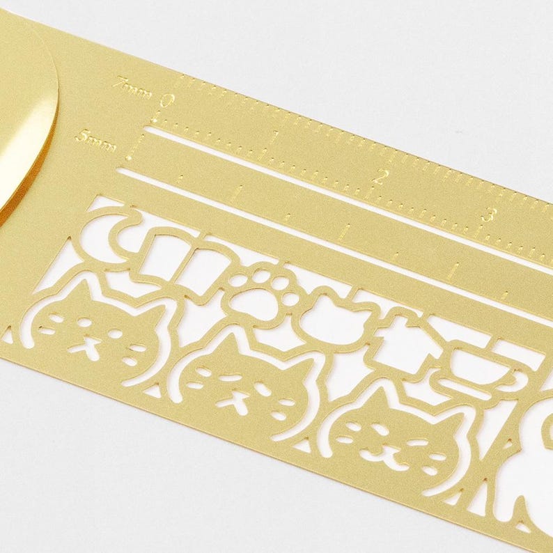 May include: A gold-coloured metal ruler featuring cut-out cat faces, paw prints, and other shapes. The ruler has measurement markings in centimetres and millimetres. Ideal for crafting and scrapbooking.