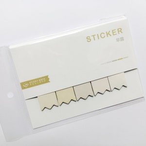 以下が含まれることがあります： 白い背景に付箋のパッケージ。パッケージには、金色の文字で「STICKER」という文字が入った白いカードが入っています。ベージュ色の旗型の付箋が5枚見えます。パッケージは透明なビニールスリーブに入っています。