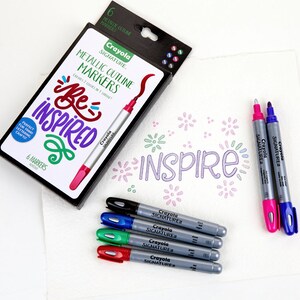 May include: A set of six metallic outline markers in a black box with the words "Crayola Signature Metallic Outline Markers" and "Be Inspired". The markers are in colors like pink, green, blue, and black. The box also includes a sample of the markers used to write the word "Inspire" on a white surface.