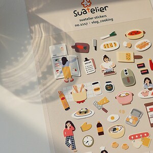 Pu&ograve; includere: Un foglio di adesivi colorati con illustrazioni di cibo, elettrodomestici da cucina e persone che cucinano. Gli adesivi sono etichettati "Suatelier stickers no.1137 : vlog_cooking".