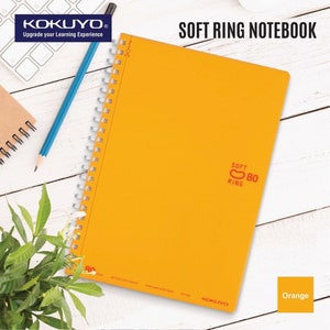 Op de afbeelding: Oranje soft ring notitieboek met 80 vellen. Het notitieboek heeft een zachte ringband en een Kokuyo-logo op de omslag. Het notitieboek ligt op een wit houten oppervlak.