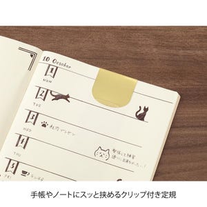 May include: Open notebook with a gold-coloured clip and cat illustrations. The page is dated October 10th, with handwritten notes and drawings. The notebook is open on a wooden surface.