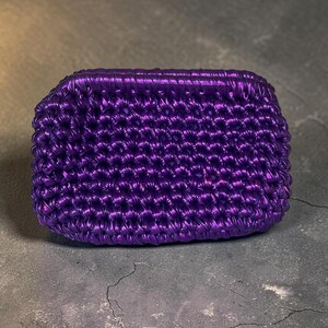 Pode incluir: Uma clutch retangular roxa metálica com uma aparência texturizada e tecida. A clutch é de cor sólida e parece ser feita de um material brilhante. A bolsa está sobre uma superfície cinza.