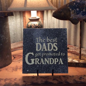 May include: A black slate sign with white text that reads "The best DADS get promoted to GRANDPA". The sign is on a small black stand and is sitting on a wooden surface. There is a lamp and a fish figurine in the background.