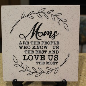 May include: White ceramic tile with a black floral wreath design and the text "Moms are the people who know us the best and love us the most."