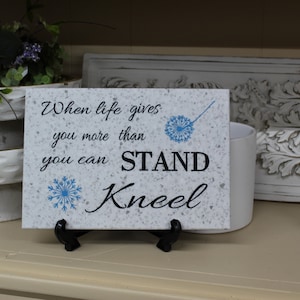 May include: White ceramic tile with a blue and black inspirational quote that reads "When life gives you more than you can stand, kneel". The tile is on a black stand and sitting on a shelf.