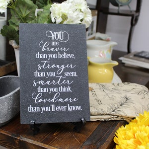 May include: A black slate sign with white text that reads "YOU are braver than you believe, stronger than you seem, smarter than you think, loved more than you'll ever know."