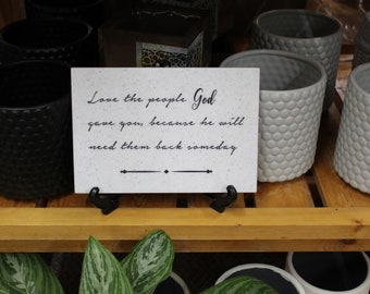 May include: A white sign with black text that reads "Love the people God gave you, because he will need them back someday". The sign is sitting on a wooden shelf with black and gray textured pots.