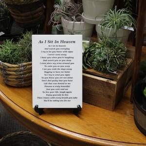 May include: A white card with a black stand sits on a wooden table. The card has a poem titled "As I Sit In Heaven" printed on it. The poem is about watching over loved ones from heaven. The table is surrounded by various potted plants.