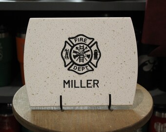 May include: A rectangular off-white stone plaque with a speckled texture, featuring a black printed fire department emblem with a ladder, helmet, and hydrant, and the name MILLER printed in black capital letters below the emblem.