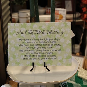 May include: A decorative card with the text "An Old Irish Blessing" in black script. The card has a light green floral background and is displayed on a white ceramic dish with a gold rim. The card is held by a black metal stand.