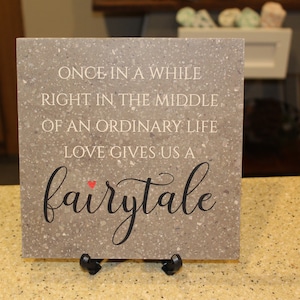 May include: A gray stone tile with a white inscription that reads "Once in a while right in the middle of an ordinary life love gives us a fairytale". A small red heart is above the word "fairytale".